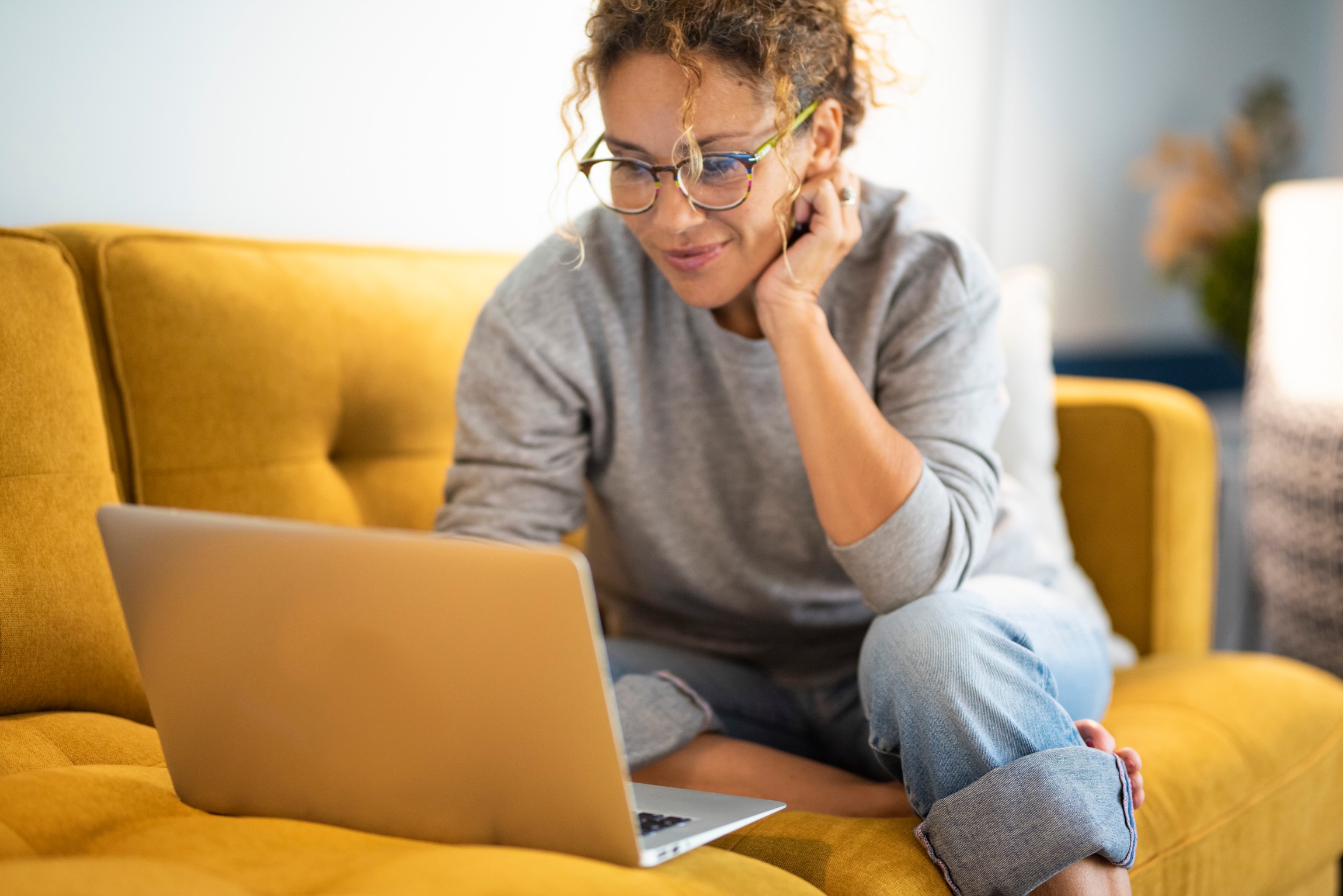 Female sitting on a yellow couch using laptop and internet connection and smile. Happy woman in indoor technology leisure activity. Social media life account concept. Adult Lady writing on computer creating passive income