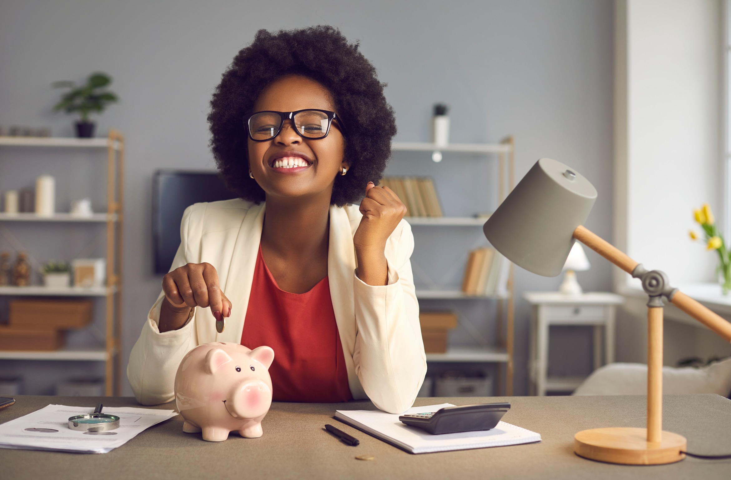 why you need to set financial goals. Lovely satisfied, excited happy young african american woman in eyewear with yes gesture putting coin into piggybank